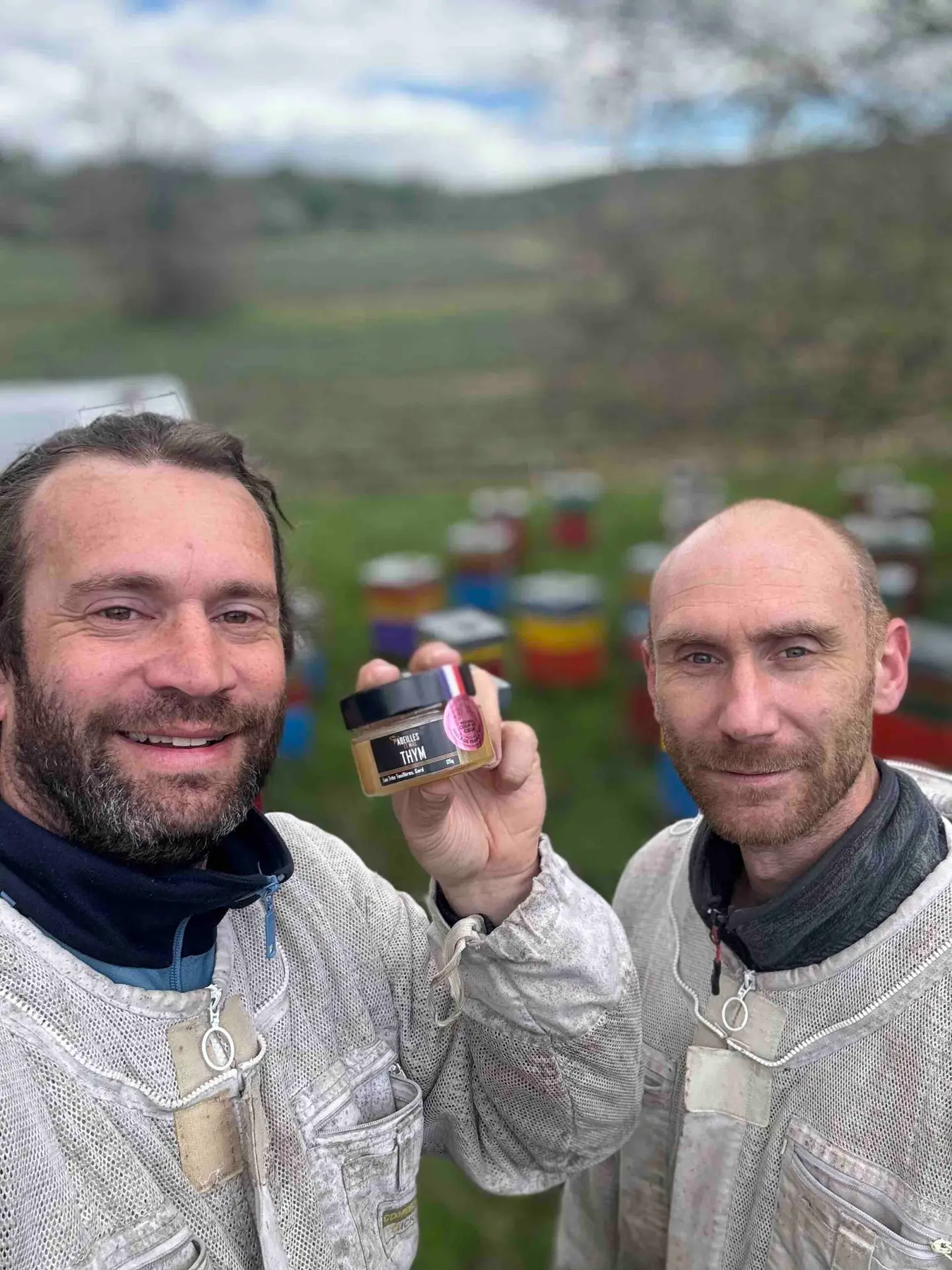 Nicolas et Rémi, apiculteurs récoltants, présentant un pot de miel de thym devant leurs ruches colorées.
